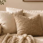 Beige sofa with textured pillows and a chunky knitted blanket draped over the arm, in a cozy living room.